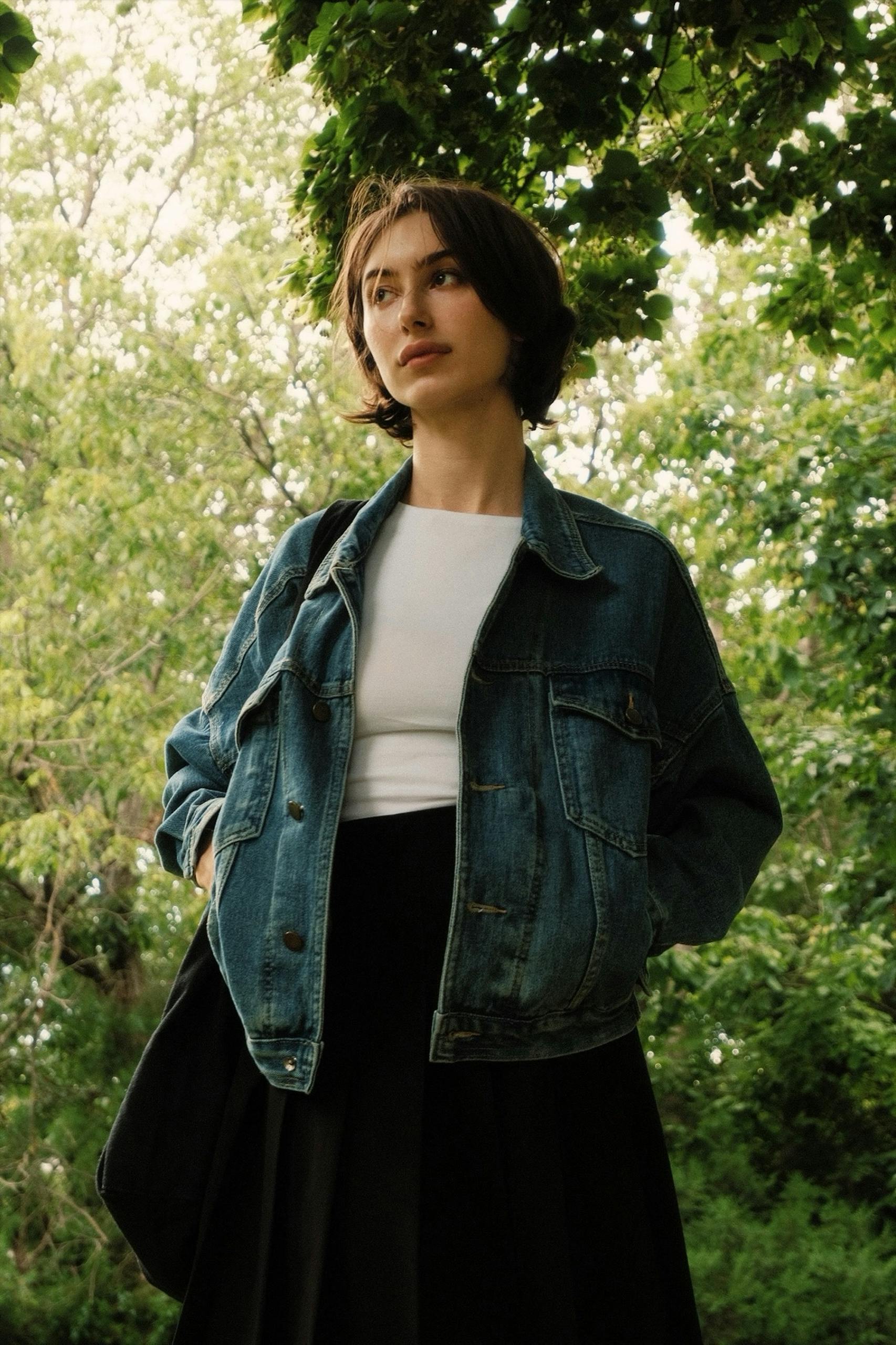 Young woman in denim jacket standing in lush green park during the day.